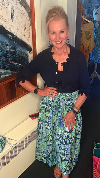 Woman wearing the Gretchen Scott tropical blue maxi skirt paired with a navy cardigan for an elegant coastal look. Versatile resort wear with hidden pockets and a flattering smocked waistband for travel and everyday summer outfits.